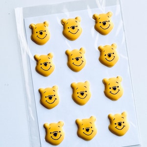 May include: A sheet of eleven yellow Winnie the Pooh character face stickers arranged in a grid pattern. Each sticker features a bright yellow bear head with black printed details for the eyes, nose, and a smiling mouth, all encased in a clear plastic sleeve.