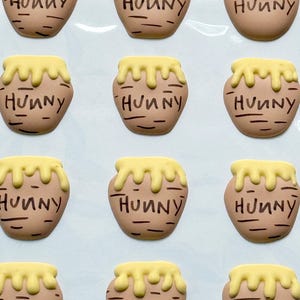 May include: A set of twelve flat, tan-colored honey pot-shaped embellishments arranged in a grid. Each pot features a yellow drip design at the top and the word "HUNNY" written in black, stylized lettering across the center of the tan surface.