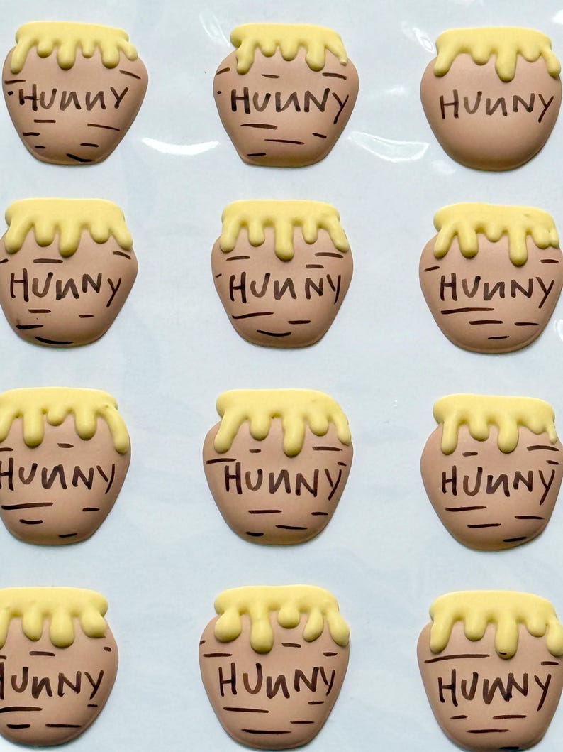May include: A set of twelve flat, tan-colored honey pot-shaped embellishments arranged in a grid. Each pot features a yellow drip design at the top and the word "HUNNY" written in black, stylized lettering across the center of the tan surface.