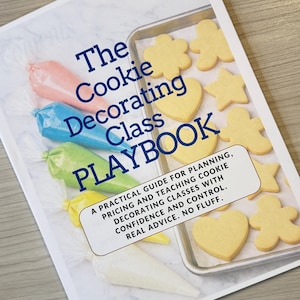 May include: A cookbook titled "The Cookie Decorating Class Playbook" with blue text. The cover shows piping bags with pink, blue, green, and yellow icing. A baking tray holds various shaped biscuits.