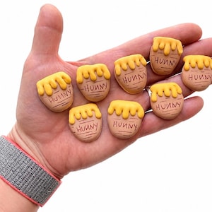 May include: Seven small, tan, pot-shaped charms with yellow, dripping honey designs on top. Each charm features the word "HUNNY" inscribed in black lettering on the front. The items are shaped like miniature honey pots with a smooth, matte finish.