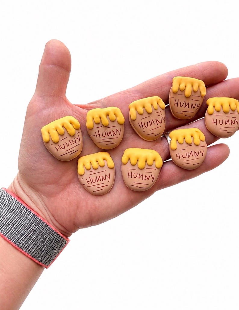 May include: Seven small, tan, pot-shaped charms with yellow, dripping honey designs on top. Each charm features the word "HUNNY" inscribed in black lettering on the front. The items are shaped like miniature honey pots with a smooth, matte finish.