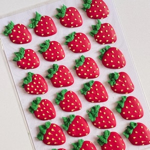 May include: A collection of twenty-one small, red strawberry-shaped decorations arranged in a grid on a white sheet. Each strawberry features a bright red body with yellow seed-like dots and a textured green leaf cluster at the top.