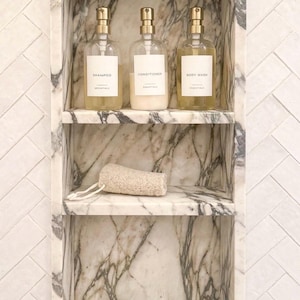 Puede incluir: Un nicho de ducha de mármol con tres estantes con vetas blancas y grises. El estante superior contiene tres botellas de vidrio transparente con dosificador y tapas doradas, etiquetadas como Shampoo, Conditioner y Body Wash. Una esponja de baño beige texturizada descansa en el estante central.