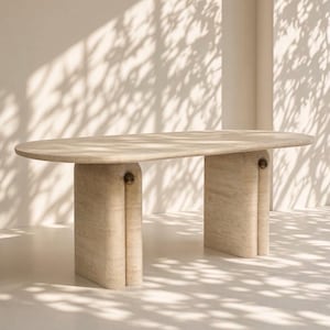 Op de afbeelding: Een ovale eettafel gemaakt van beige travertijnsteen. De tafel heeft een glad, plat blad dat wordt ondersteund door twee dikke, gebogen voetstukken. Elk voetstuk is voorzien van een klein, rond metalen detail op het bovenste verbindingspunt.