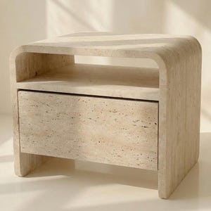 May include: A beige travertine nightstand featuring a curved, waterfall-style frame with a single rectangular drawer and an open storage shelf above. The natural stone surface displays a porous, textured pattern in light tan and cream tones.