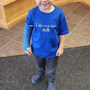 Può includere: Un bambino piccolo che indossa una maglietta blu con il testo "I do my own stunts" in bianco. Indossa pantaloni grigi e scarpe blu. Ha un gesso blu sul braccio sinistro.