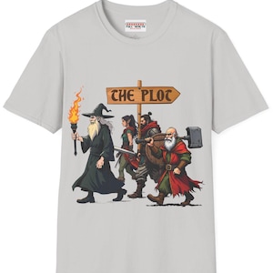 May include: Light gray t-shirt featuring a fantasy-themed illustration. The design depicts a wizard with a torch, followed by a warrior, a woman with a sword, and a dwarf with a hammer. A wooden sign reads "THE PLOT".