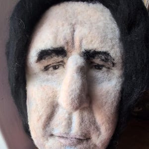 May include: A felted sculpture of a face with black hair and a pale complexion. The sculpture has detailed facial features, including eyebrows, eyes, a nose, and a mouth. The sculpture is likely a representation of a person.