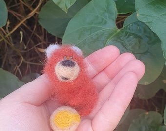 Pode incluir: Um pequeno panda vermelho de feltro feito à mão com orelhas brancas e uma bola amarela. O panda está sentado em uma mão.
