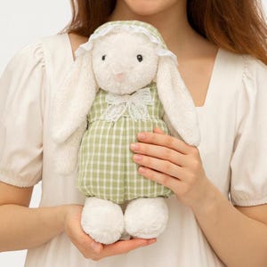 Puede incluir: Un peluche de conejo blanco con un vestido a cuadros verdes y blancos con cuello y gorro de encaje blanco. El conejo tiene orejas largas y caídas y un pelaje suave y esponjoso. El juguete está diseñado para ser abrazado y está en las manos de una persona.