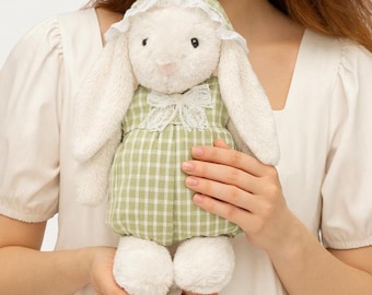 Pode incluir: Um coelho de pelúcia branco vestindo um vestido xadrez verde e branco com gola e touca de renda brancas. O coelho tem orelhas longas e caídas e pele macia e fofa. O brinquedo foi projetado para ser aconchegante e está nas mãos de uma pessoa.