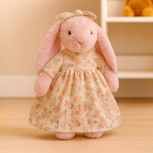 May include: A plush pink bunny wearing a floral print dress and bow. The bunny has long, floppy ears and a sweet expression. The dress features a pattern of pink roses and green leaves. The toy is standing on a light-colored wooden floor.