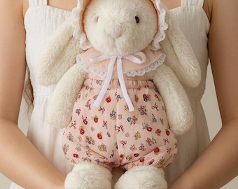 Pode incluir: Uma boneca de coelho branco de pelúcia usando um capuz rosa e bloomers estampados. O coelho tem orelhas longas e pés fofos. A boneca é segurada em frente a um vestido branco.