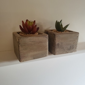 Puede incluir: Dos pequeñas macetas de madera con suculentas artificiales. Las macetas son de color marrón claro con un acabado desgastado. Las suculentas son de color rojo y verde.