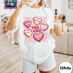 May include: White t-shirt with a graphic of pink candy hearts with the words "Loved", "Strong", "Kind", "Creative", and "Important". The words "You Are" are in the center. The shirt is paired with gray shorts.
