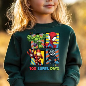 A young girl wearing a green sweatshirt with a graphic of four cartoon characters: the Hulk, Spider-Man, Iron Man, and Black Widow, along with the text "100 SUPER DAYS".