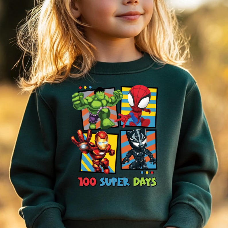 A young girl wearing a green sweatshirt with a graphic of four cartoon characters: the Hulk, Spider-Man, Iron Man, and Black Widow, along with the text "100 SUPER DAYS".
