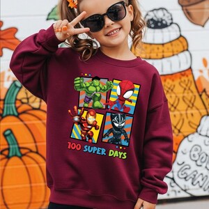 A young girl wearing a maroon sweatshirt with a graphic of four superhero characters and the text "100 SUPER DAYS" is smiling and making a peace sign.