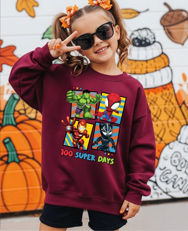 A young girl wearing a maroon sweatshirt with a graphic of four superhero characters and the text "100 SUPER DAYS" is smiling and making a peace sign.