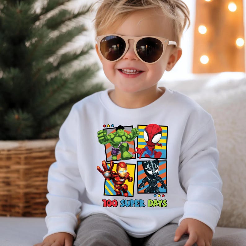 A young child wearing a white sweater with a graphic of four cartoon characters - Hulk, Spider-Man, Iron Man, and Black Widow - smiling and posing for the camera.
