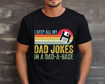 May include: Black t-shirt with a retro design. The shirt features the text "I KEEP ALL MY DAD JOKES IN A DAD-A-BASE" in a vintage font, with a floppy disk graphic and a sunset-style background.
