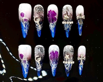 May include: A set of ten long, pointed artificial nails. Each nail features a design with white, purple, and blue hues. Some nails have lace patterns, floral accents, or silver embellishments. The nails are arranged on a black surface.