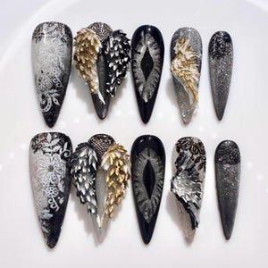 May include: A collection of ten long, pointed artificial nails. The nails showcase diverse designs, including silver glitter, black lace patterns, and 3D wing accents in gold and silver. The nails are displayed on a white surface.