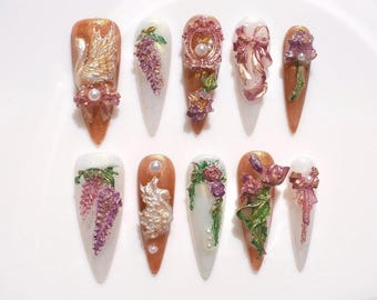 May include: Ten long, almond-shaped artificial nails are displayed. Each nail showcases detailed 3D designs, such as flowers, bows, and ornate frames, in gold, white, and purple hues. Some nails are adorned with pearl accents.