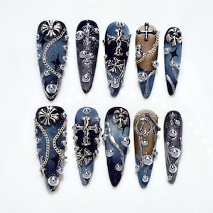Op de afbeelding: Een set van tien lange, amandelvormige kunstnagels. Elke nagel heeft een donkerblauw en zilver design met versieringen zoals kruisen, kettingen en strass-steentjes. De nagels hebben een gotische of punk esthetiek.