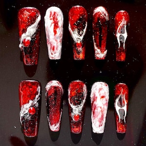 Puede incluir: Un conjunto de diez uñas postizas en forma de ataúd. Cada uña es de color rojo intenso con un acabado brillante, con diseños abstractos blancos y plateados. Algunas uñas tienen un pequeño acento rojo. Las uñas están dispuestas sobre una superficie oscura.