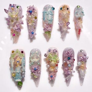 Op de afbeelding: Een reeks van tien lange, puntige kunstnagels, elk met een uniek ontwerp. De nagels zijn versierd met ingewikkelde decoraties in roze, paars, blauw en goud, met bloemen, feeën en juwelen. Ze zijn gerangschikt op een wit oppervlak.