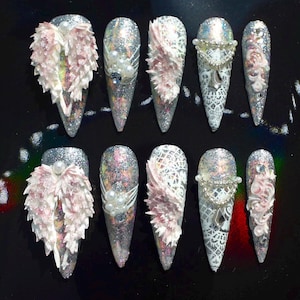 May include: A set of ten long, pointed artificial nails. Each nail is decorated with glitter, pearls, lace, and 3D angel wing designs in shades of pink, silver, and white. The nails are arranged on a black surface.