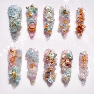Peut inclure: Dix faux ongles de forme amande allongée. Chaque ongle est décoré de manière unique avec des ornements bleus clairs, dorés et roses, notamment des cristaux, des perles et des motifs floraux. Les ongles sont disposés sur une surface blanche.