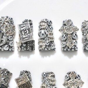 Puede incluir: Una colecci&oacute;n de dijes de u&ntilde;as plateados con varios dise&ntilde;os, incluyendo corazones, billetes de d&oacute;lar, cruces y elementos decorativos. Los dijes est&aacute;n adornados con peque&ntilde;as cuentas brillantes y detalles intrincados.