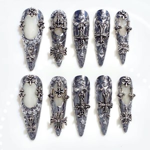 May include: A collection of ten silver-coloured nail covers with intricate designs. Each cover has a pointed shape, adorned with crosses, floral patterns, and clear gemstones. The nail covers are displayed on a white surface.