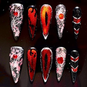 May include: A set of ten long, pointed false nails. Designs include red, black, and white patterns, glitter, and red gem accents. Some nails feature a red eye design, while others have a floral pattern.