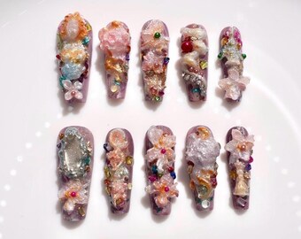 May include: A collection of ten long, purple artificial nails, each adorned with intricate, three-dimensional floral and gem designs. The nail art features a variety of colors, including pink, orange, white, and green, creating a glamorous and artistic aesthetic.
