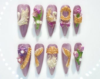 May include: Ten lavender-coloured artificial nails, each decorated with detailed 3D embellishments. Designs include swans, flowers, bows, and ornate frames in gold, white, and pink. The nails are arranged on a white surface.