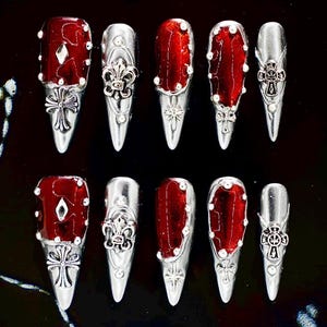 May include: A set of ten long, pointed false nails. Each nail has a deep red, glossy finish with silver-coloured embellishments. Designs include crosses, fleur-de-lis, and geometric shapes, creating a gothic or edgy aesthetic.