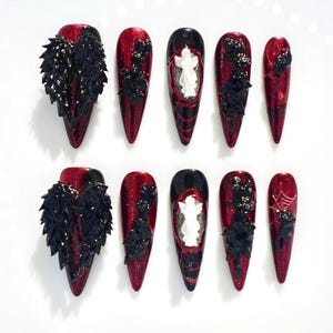 Puede incluir: Un conjunto de diez uñas artificiales largas y puntiagudas. Las uñas presentan una combinación de colores rojo y negro con detalles de purpurina. Algunas uñas tienen adornos negros en forma de pluma, otras diseños de fantasmas blancos o telarañas.