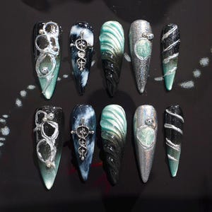 May include: A collection of ten long, pointed artificial nails. Each nail features unique designs, including silver embellishments, swirling patterns, and gradient color schemes of black, silver, and teal. The nails are arranged on a black surface.