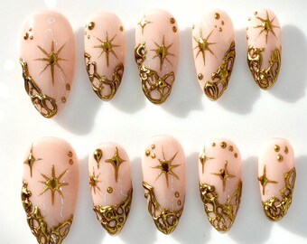 以下が含まれることがあります： 淡いピンク色のベースに、金色の星のデザインとベースに金色の透かし模様が施された10本のつけ爪。小さな金色のドットも付いており、天体テーマを演出しています。