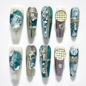 May include: A set of ten artificial nails with various designs. Some nails feature white lace, teal accents, and silver embellishments. Others have a grid-like pattern or metallic finishes. The nails are arranged on a white surface.