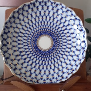 May include: A white porcelain plate with a blue and gold geometric pattern. The plate has a scalloped edge and is displayed on a wooden stand.