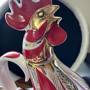 May include: A red and gold ceramic rooster figurine with white accents. The rooster is depicted with its head tilted back and its beak open, as if it is crowing. The figurine is detailed with gold paint and has a white base.