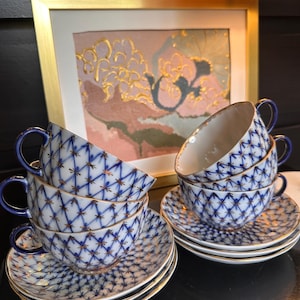 May include: A set of six teacups and saucers. The cups and saucers are white with a blue lattice pattern and gold trim. The cups have blue handles. The background includes a framed painting with gold accents.