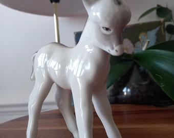 May include: A ceramic figurine of a donkey in a standing pose. The donkey is primarily white with gray accents on its ears and back. The figurine has a glossy finish and is set on a wooden surface.
