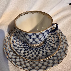 Può includere: Servizio da tè in porcellana composto da tazza, piattino e piattino da dessert. Il set ha una base bianca con un motivo a griglia blu e bordi dorati. La tazza ha un manico blu scuro e bordi smerlati.