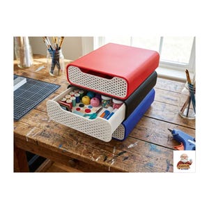May include: Three-tier craft storage drawers in red, black, and blue. The top drawer is red, the middle is black, and the bottom is blue. The open white drawer reveals craft supplies like spools of thread, buttons, and scissors. The drawers are on a wooden table.